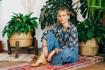 Young woman in jeans and a blouse sitting on the floor next to the foliage of tropical plants.