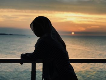 Silhouette woman standing by railing against sea during sunset