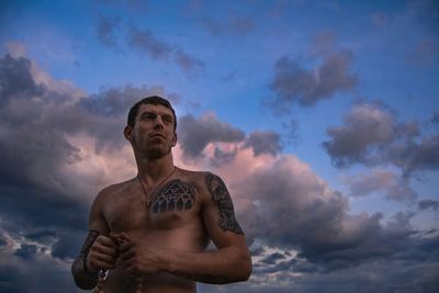 Low angle view of shirtless man with tattoo against cloudy sky