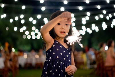 Portrait of girl holding sparkler while standing against lights during night