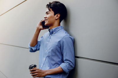 Young man using mobile phone while standing against wall