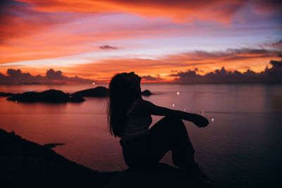 Silhouette man sitting by sea against orange sky