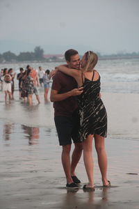 Friends embracing while standing on shore at beach against sky