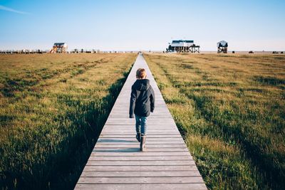 Rear view of boy walking on boardwalk over grassy field