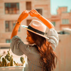 Woman holding hat standing against built structure