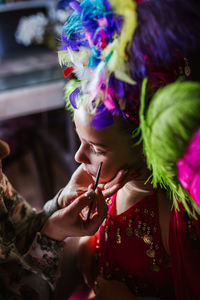 Crop unrecognizable side view of woman hand applying eyeliner on eye of cute little girl with closed eyes in colorful feather headgear costume during carnival preparation at home