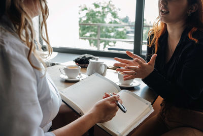 Midsection of woman writing on diary while having discussion with colleague at cafe