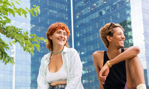 Positive multiracial friends in casual wear sitting together on border near modern building in city on summer day looking away