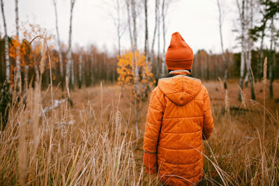 Pensive girl in orange coat stands in forest among dry grass and golden trees, outdoor, fall vibes