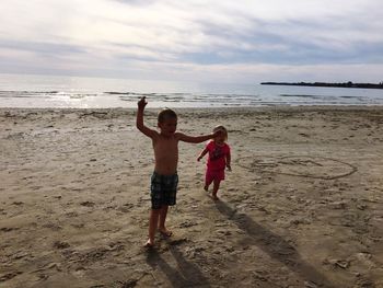 Full length of cute siblings playing on sand at beach against sky