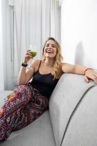 Young blonde woman sitting on the sofa drinking a vegetable smoothie