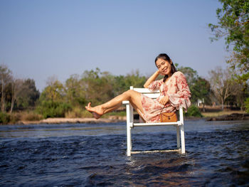 Woman sitting on chair at riverbank against clear sky