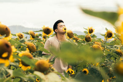 Young man with closed eyes standing amidst sunflowers against sky