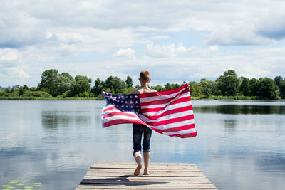 Rear view of young man holding american flag while running on pier against sky