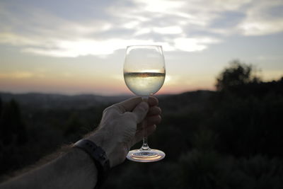 Close-up of hand holding wineglass against sky during sunset