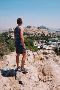 Rear view of man standing on rock formation while looking at town against sky