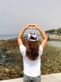Rear view of girl making heart shape with hands against sky