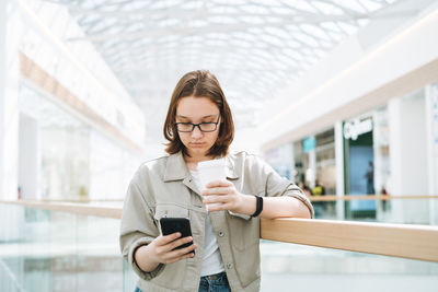 Young brunette teenager girl student in glasses using mobile with paper coffee cup at shopping mall