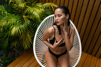 Portrait of a young woman sitting on white wicker chair on wooden floor in front of palm trees