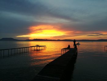 Silhouette friends at pier by lake against cloudy sky during sunset