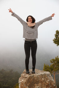 Full length of carefree young woman with arms raised standing on rock during foggy weather