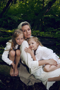 Family blonde mother with two children in a white dress is lying on tree log in the forest barefoot
