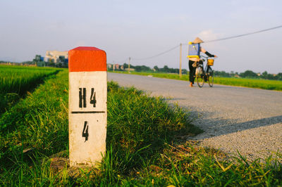 Sign on grass against woman with bicycle on road