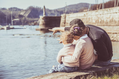 Rear view of man sitting with daughters by lake