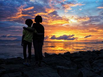 Rear view of siblings standing on rock at beach during sunset
