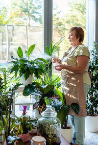 Adult senior retrired woman taking care of plants at home spraying water at flowerpot in living room