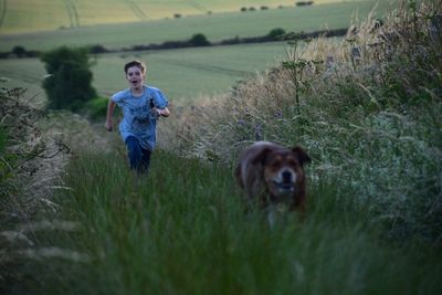 Boy running behind dog amidst grassy field