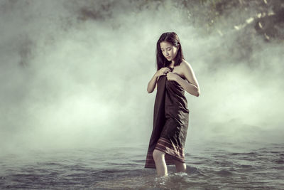 Young woman wrapped in textile standing in river during foggy weather