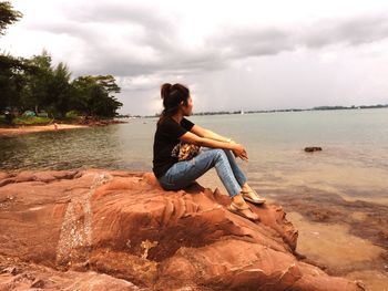 Young woman sitting on rock by sea against sky