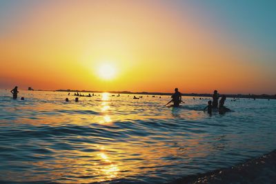 Silhouette people in sea against orange sky