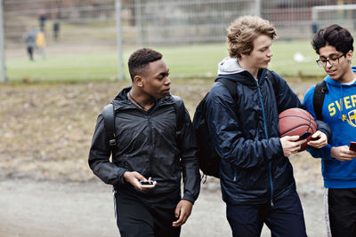 Multi-ethnic friends talking while holding mobile phones on street against playing field