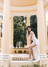 A beautiful brunette lady in an elegant wedding dress poses among the columns in the old city park