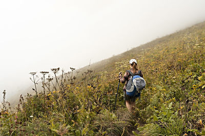Rear view of young woman with big backpack trekking poles walking along mountain trail hiking in fog