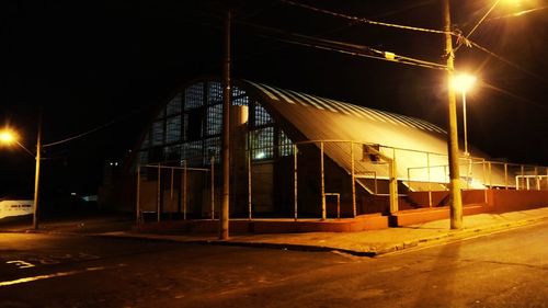 Illuminated street by building against sky at night