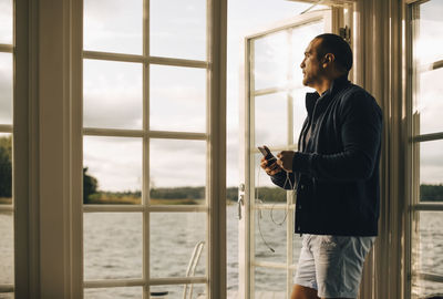 Thoughtful mature man using mobile phone while standing at doorway in holiday villa