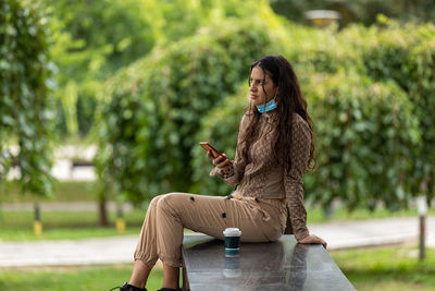 Attractive teen girl with smartphone and coffee sitting in a park and having a phone conversation. 
