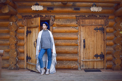 Woman in a blue hat and sweater stands at the door of a wooden house made of logs in autumn