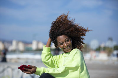 Smiling young woman using smart phone while sitting against sky