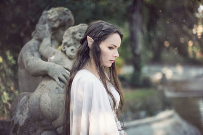 Side view of thoughtful fairy elf young woman standing by sculptures