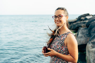 Portrait of smiling young woman standing by sea against sky