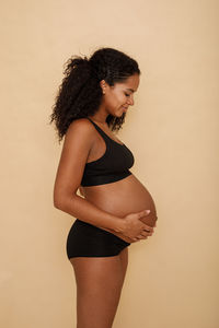 Side view of pregnant woman with hands on stomach standing against beige background