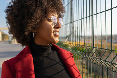 Side view of african american female with afro hairstyle and wearing eyeglasses and formal crimson jacket looking away through metal grid of fence in sunlight