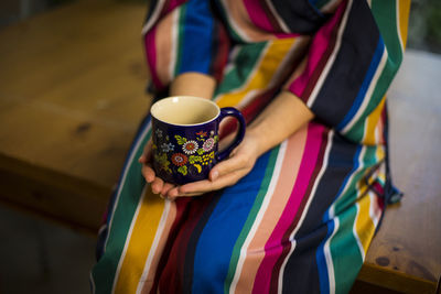 Woman in multi colored clothing holding coffee cup while sitting on table at home