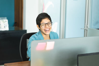 Cheerful hispanic female entrepreneur with short hair in stylish outfit sitting at table with laptop and laughing while working in light office in costa rica in daytime