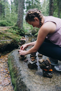 Side view of woman sitting on rock in forest stacking stones 