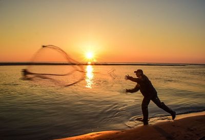 Fisherman throwing net in sea against orange sky during sunset